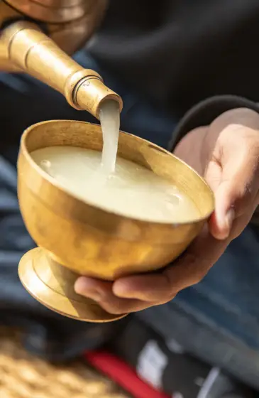 A traditional brass vessel pouring Chhaang, a fermented millet drink, into a brass bowl, symbolizing the base for crafting Raksi.